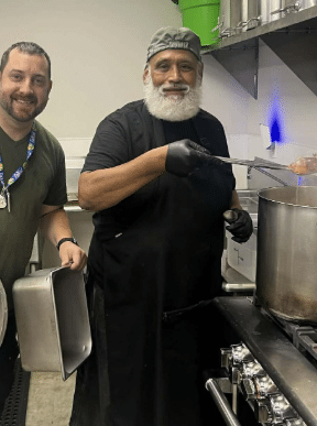 Two Cafe cooks pose for the camera. One ladles soup from the pot on the stove to show the camera.